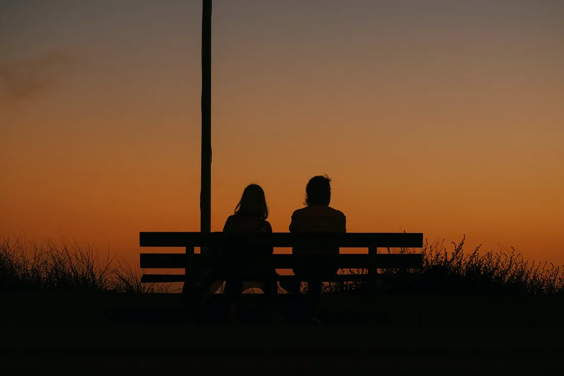 Deux silhouettes assises sur un banc devant un ciel orangé de fin de journée